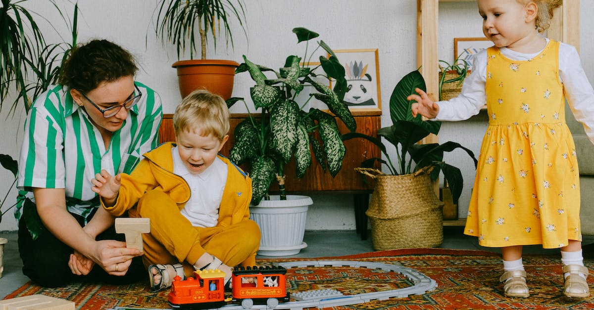 Mom and adorable little brother and sister in casual wear gathering in cozy living room during weekend and having fun together while playing with plastic railway
