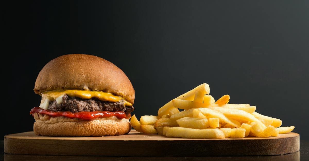 Appetizing burger with meat patty ketchup and cheese placed on wooden table with crispy french fries against black background