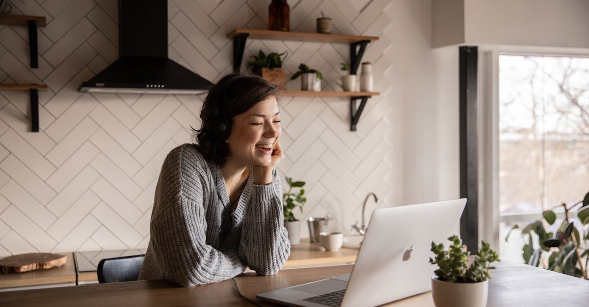 Young cheerful female smiling and talking via laptop while sitting at wooden table in cozy kitchen