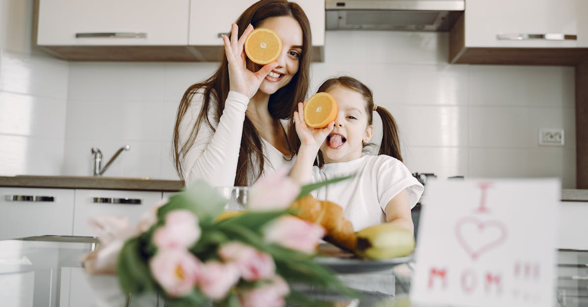 From below of cheerful mother and daughter in domestic clothes smiling and playing with oranges while sitting together at table with bouquet of tulip and drawing of child