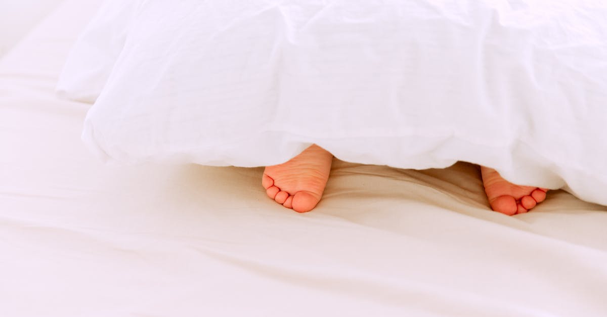 Crop tiny feet of little child lying under white blanket on comfortable bed in light bedroom at home