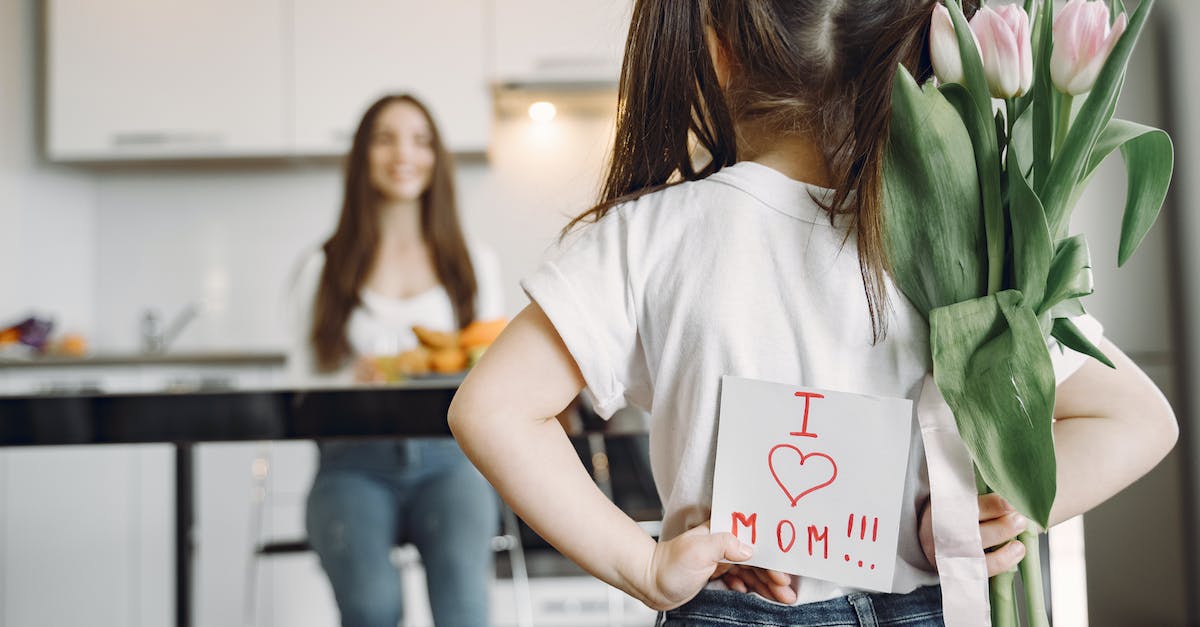 Back view of little girl with ponytails in casual clothes standing against mother with hands behind back and holding bouquet of tulips and drawing I love mom on kitchen