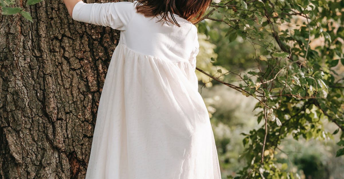 Back view full body of faceless little girl climbing on tree with green foliage while spending time in park