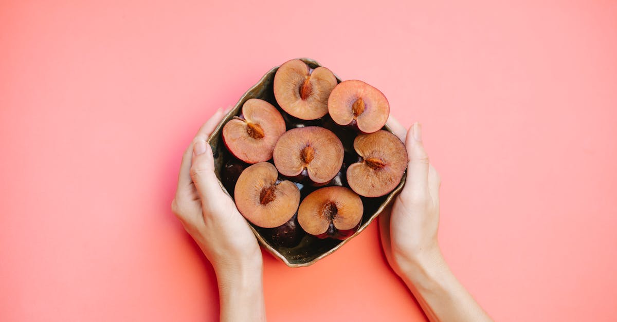 Crop faceless lady demonstrating bowl of ripe halved plums against pink background